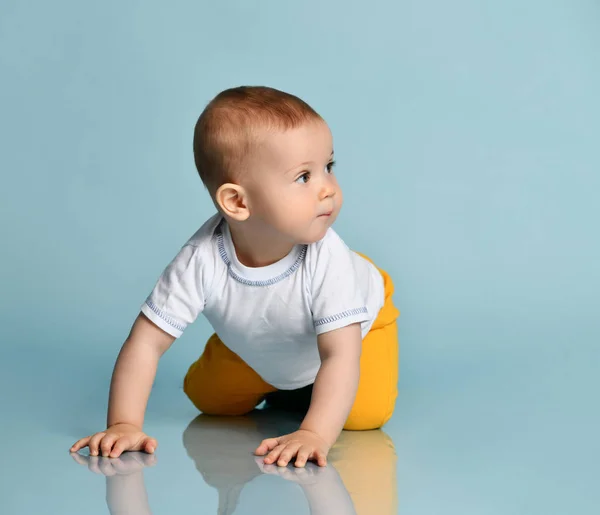 Säugling Baby Junge Kind in gelber Hose und weißem T-Shirt krabbelt auf allen Vieren und schaut beiseite auf blauem Hintergrund — Stockfoto