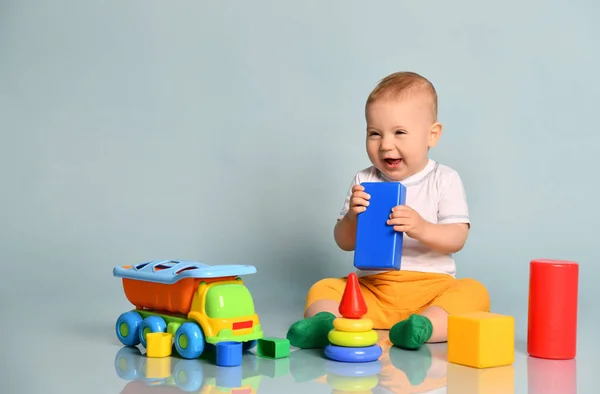 Cute uśmiechnięty śmiech niemowlę chłopiec maluch w żółtych spodniach siedzi w otoczeniu zabawek, trzymając blokadę na ustach — Zdjęcie stockowe