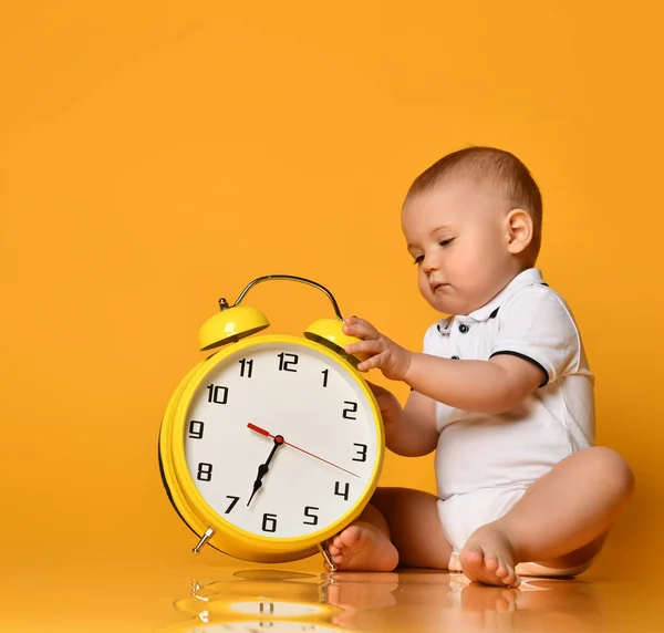 Малыш-младенец занят тем, что сидит у большого желтого будильника с руками, играющими на желтом — стоковое фото