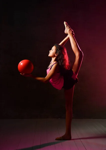 Νεαρή αθλήτρια γυμνάστρια που κάνει γυμναστικές ασκήσεις με κόκκινη μπάλα και εκτελεί κάθετες σχισμές που εκτείνονται στο σκοτάδι — Φωτογραφία Αρχείου