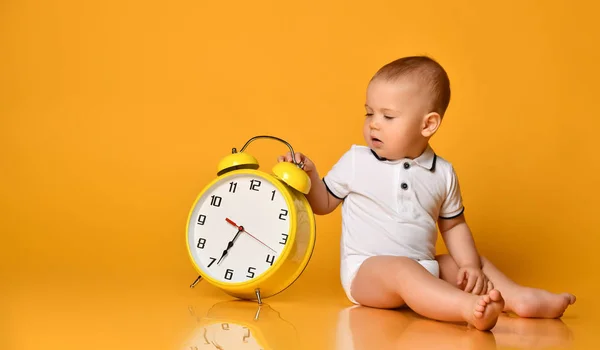 Baby boy toddler is occupied with sitting at big yellow alarm clock with hands playing showing on yellow with copy space — Stock Photo, Image