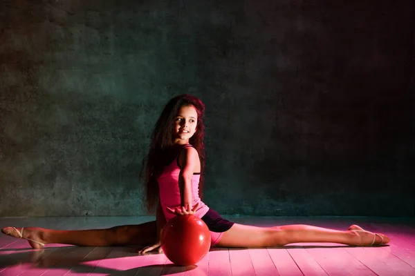 Uzun kıvırcık saçlı, genç ve zarif bir kız. Jimnastik yapıyor. Top yerde oturuyor, esniyor. — Stok fotoğraf