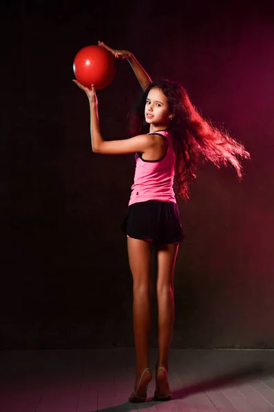 Athletic girl gymnast with long hair posing with her back to us doing exercises with a red ball holding it up, turning — Stock Photo, Image