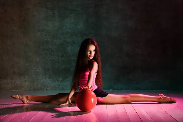 Flexible teen girl with long hair teen doing rhythmic gymnastic exercises with red ball sitting on floor stretching