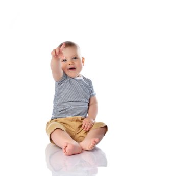 Happy infant baby boy in t-shirt and pants is sitting on the floor pointing at something up on white with copy space clipart