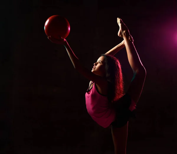 Молода дівчина робить гімнастичні вправи, виконуючи вертикальні розгалуження, що розтягуються, тримаючи червоний м'яч на довжині рук на темряві — стокове фото