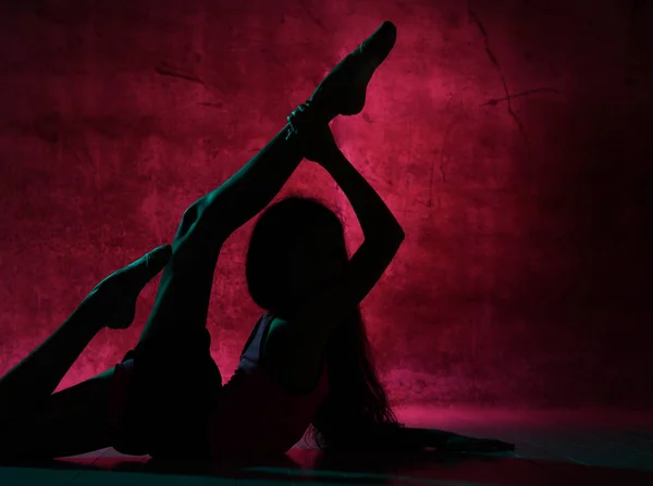 Silhouette of young slim girl doing gymnastic exercises lying on the floor stretching bent in lumbar holding legs up — 스톡 사진