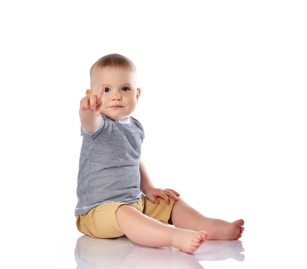 Infant baby boy in t-shirt and pants is sitting on the floor pointing at something up, pushing on white with copy space — Stock Photo, Image