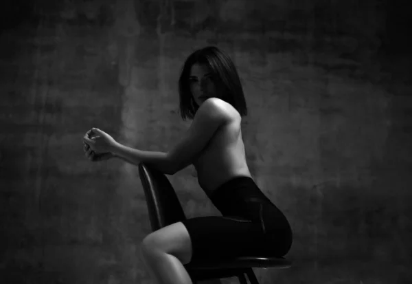 Joven mujer morena delgada en pantalones cortos deportivos negros medio desnuda sentada en la silla y mirando a la cámara sobre el fondo oscuro de la pared —  Fotos de Stock
