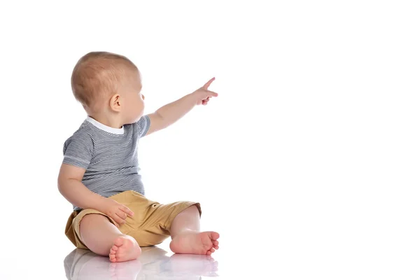 Infant baby boy in t-shirt and pants is sitting on the floor looking back and pointing at upper corner behind him — Stock Photo, Image