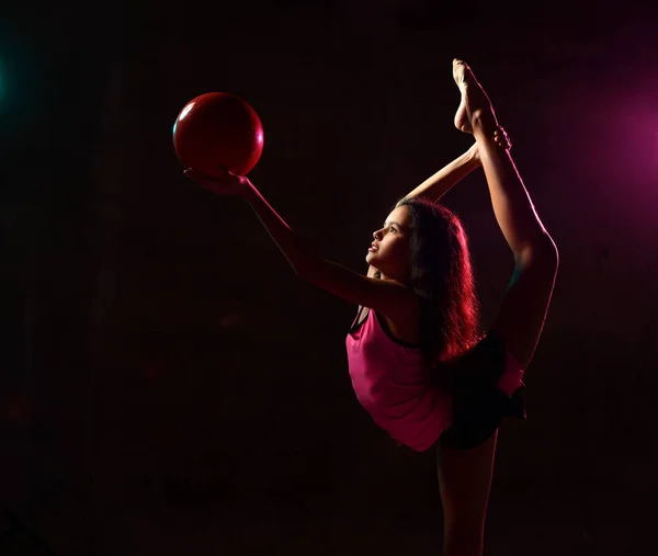 Menina fazendo exercícios de ginástica realizando divisões verticais alongamento segurando bola vermelha olhando para ele no escuro — Fotografia de Stock