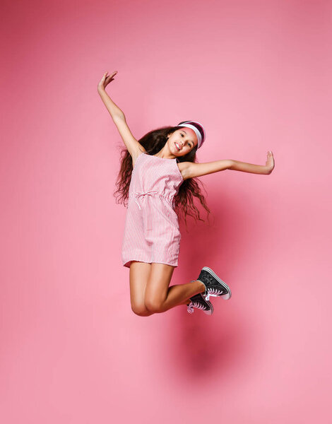 Счастливая девушка в розовых полосатых платьях прыгает высоко с поднятыми руками на розовом фоне

