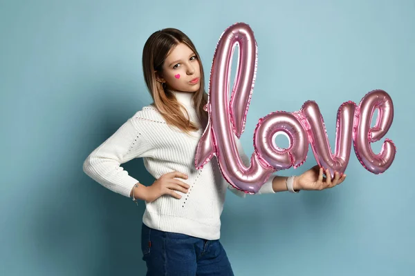 Blonde teen girl in white sweater and blue jeans holds love pink balloon on valentine day. Love word balloon — 스톡 사진