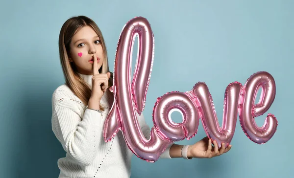 Blondes Teenie-Mädchen hält am Valentinstag einen rosafarbenen Luftballon in der Hand, der eine Geste des Schweigens zeigt. Liebe Wort Luftballons — Stockfoto