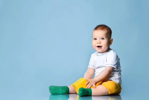 Leende spädbarn pojke småbarn i gula byxor sitter på golvet håller leka med gul kub — Stockfoto