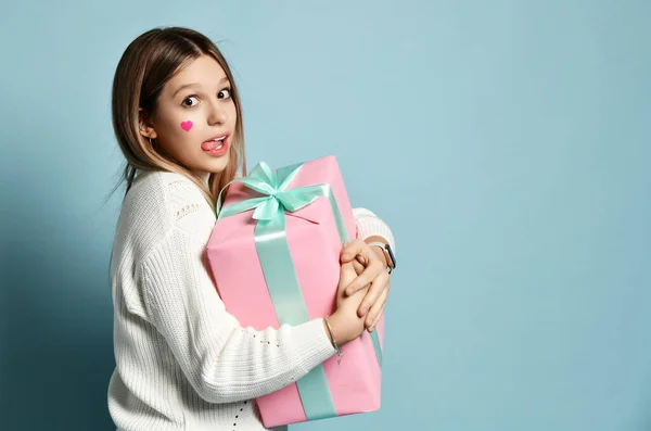 Happy young girl celebrates valentines day hugging a big pink gifts box for birthday looks at us biting her tongue — Stok fotoğraf