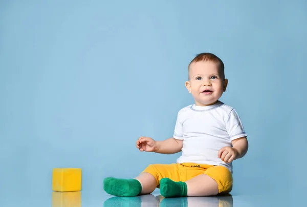 Bébé garçon tout-petit souriant en pantalon jaune est assis sur le sol près du cube jaune, regardant vers le haut — Photo