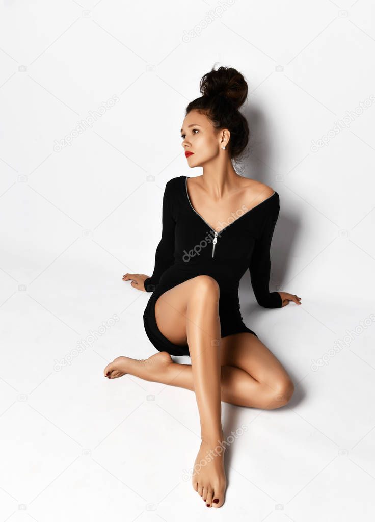 Gorgeous brunette woman in stylish short black dress is sitting against the wall leaning on her hands looking at aside