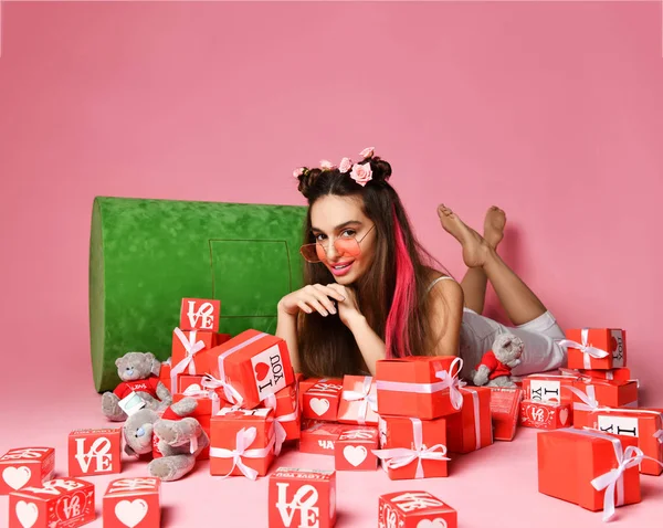Young brunette woman with a romantic hairstyle is lying on floor surrounded by boxes gifts with hearts and love on pink — Stock Photo, Image