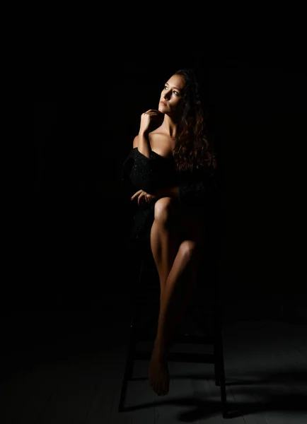 性感的女人，一头长长的卷曲的黑发，穿着黑肩裙，在黑暗中坐在高高的椅子上 — 图库照片