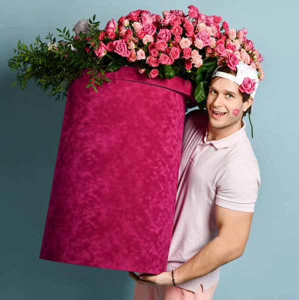 Smiling man delivery guy with rose at ear and lipstick kiss on his cheek is holding huge valentines day box with flowers