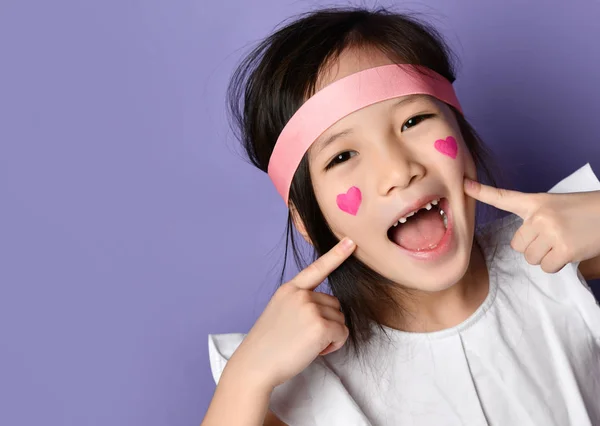 Портрет счастливой улыбчивой веселой азиата корейской девочки смеющейся указывая пальцами на щеки с красными сердцами знак на щеке — стоковое фото