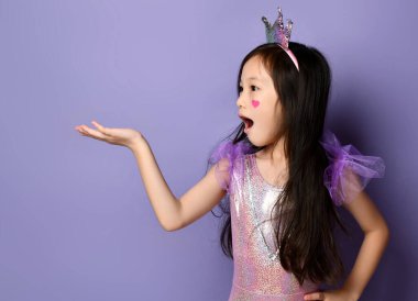 Surprised asian kid girl princess in party dress and crown is holding something on her open palm looking at it