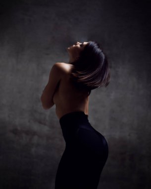 Young slim brunette woman in black sport shorts and topless is standing backwards with naked back over dark background