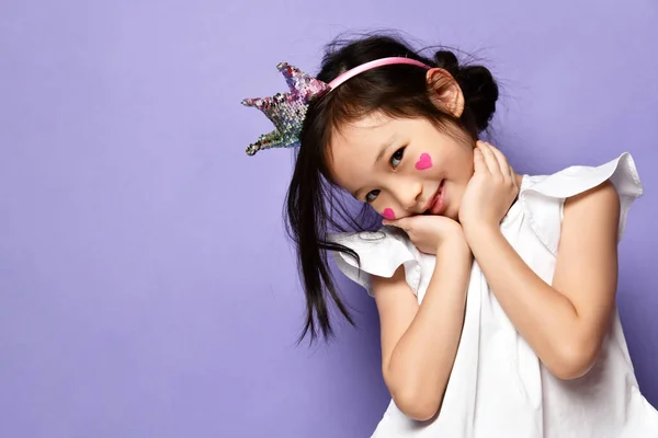 Carino asiatico coreano bambino ragazza in corona e con rosso cuori segno su il suo guancia copre guance con il suo mani e sorrisi — Foto Stock