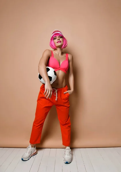 Cool atletická žena s růžovými vlasy a stylové hledí v jasném sportovním oblečení je pózování s fotbalovým míčem pod paží — Stock fotografie