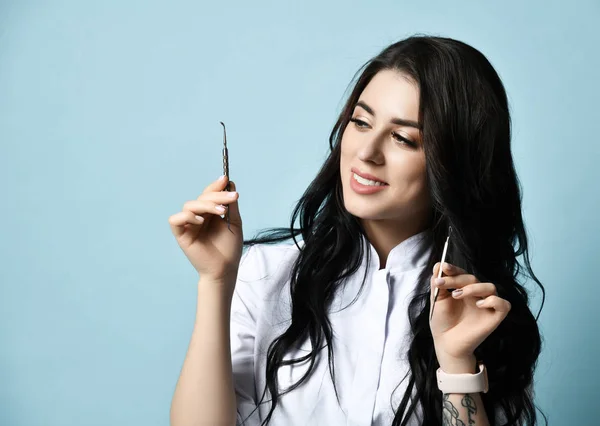 Young smiling sexy brunette woman in white medical uniform is holding cosmetology tools over blue background — 스톡 사진