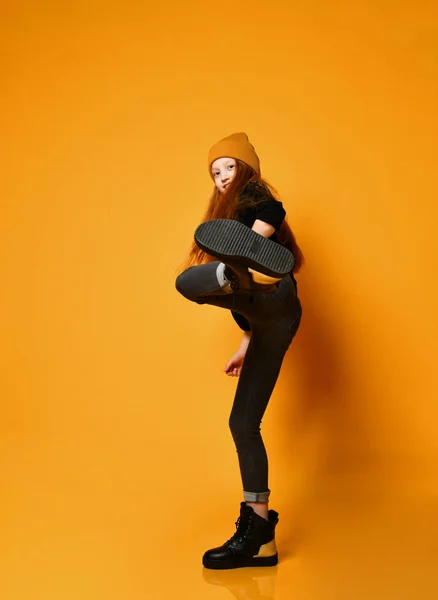 Smiling frolic red-haired girl in hat black tight jeans and boots is kicking over orange background — Φωτογραφία Αρχείου
