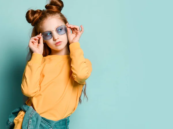 Cooles rothaariges Teenie-Mädchen in gelbem Sweatshirt probiert eine stylische blaue Brille an und schaut beiseite auf freien Kopierplatz — Stockfoto