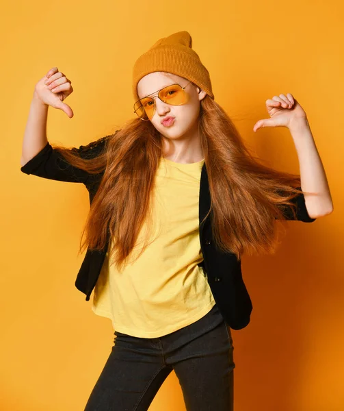 Крута дівчина-підліток з довгим рудим волоссям у в'язаному капелюсі, жовті окуляри та футболка вказує пальці на себе — стокове фото