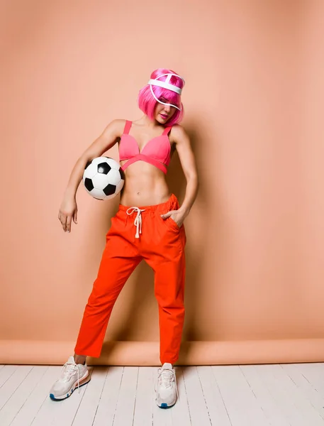 Mulher atlética legal com cabelo rosa e viseira elegante em sportswear brilhante está posando com bola de futebol sob seu braço — Fotografia de Stock