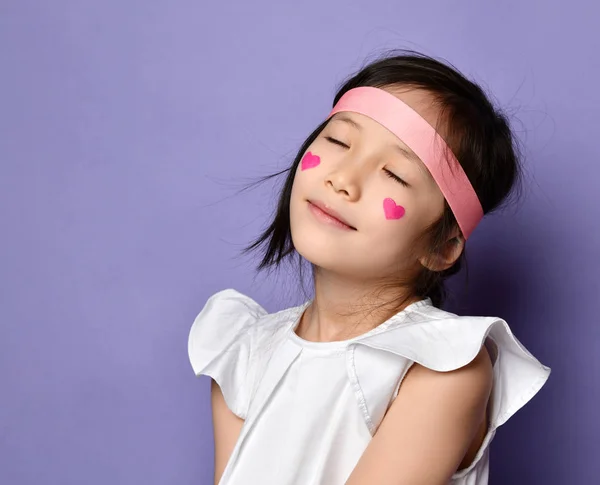 Portrait of asian Korean kid little girl with red hearts sign on cheek posing with closed eyes feeling bliss — стокове фото