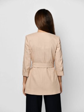 Young brunette woman is posing backwards in stylish beige formal suit jacket with cloth belt