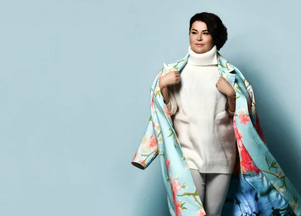 Brunette girl in white sweater and jeans, coat with floral print. Smiling, holding coat with her hands, posing on blue background — Stok fotoğraf