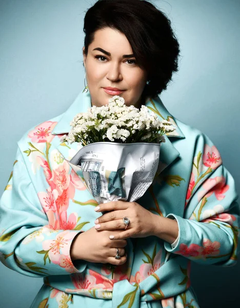 Жінка в пальто з квітковим принтом. Усміхнений, тримає букет, загорнутий в папір з проектом квартири, позує на синьому фоні — стокове фото