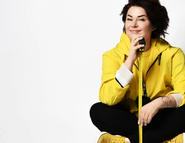 Modelo em t-shirt amarela, ténis e casaco, calças pretas. Mostrando fita métrica, posando sentado no chão, isolado no branco — Fotografia de Stock