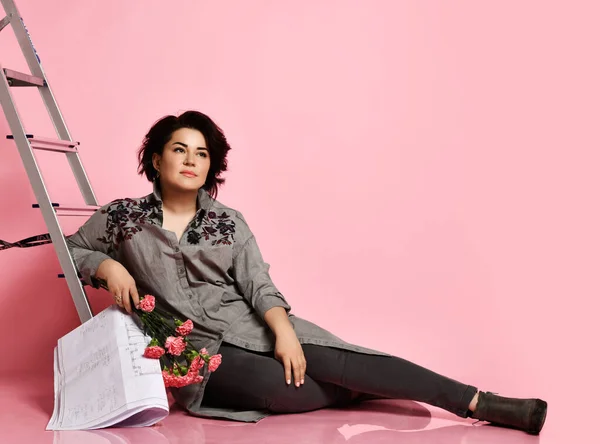 Жінка в сірій сорочці, штани та черевики. Сидить біля сходів, тримає букет і малює будинок, позує на рожевому фоні — стокове фото