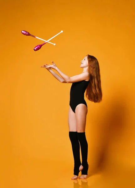 Smiling teen girl gymnast with long red hair in swimsuit trows gymnastic clubs up standing tiptoe — Stok fotoğraf