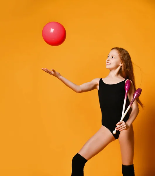 Redhead girl gymnast in black swimsuit with rhythmic gymnastic clubs at her shoulder is looking at ball she throws up — Stok fotoğraf