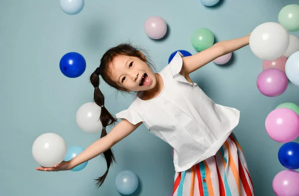 Joyful asian kid girl with long black plait is having fun surrounded by colorful air balloons, spread hands — Φωτογραφία Αρχείου