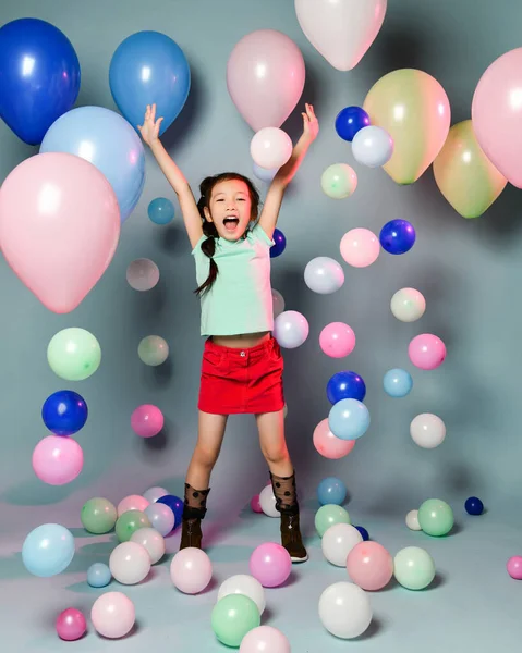 Glückliches asiatisches Mädchen in weißem T-Shirt und rotem Rock hat Spaß, umgeben von vielen bunten Luftballons, die die Hände in die Höhe halten — Stockfoto