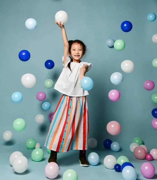 Ευτυχισμένο κορίτσι ασιατικό παιδί σε κομψό ριγέ maxi φούστα έχει διασκέδαση παίζει περιβάλλεται από πολύχρωμα μπαλόνια αέρα κρατώντας εμετό — Φωτογραφία Αρχείου