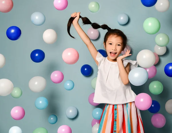 Веселый резвый азиатский ребенок девочка весело в окружении красочных воздушных шаров держа ее длинные черные косы — стоковое фото