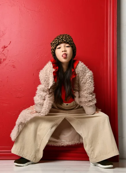 Frolic asian kid girl in beige furcoat, wide velvet pants is posing with hands on her knees sticking out her tongue — Stok fotoğraf