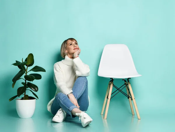 Dame im Pullover, Jeans, Turnschuhen. Lächelnd, ihr Gesicht stützend, auf dem Boden zwischen Stuhl und Ficus sitzend, posierend auf blauem Hintergrund — Stockfoto