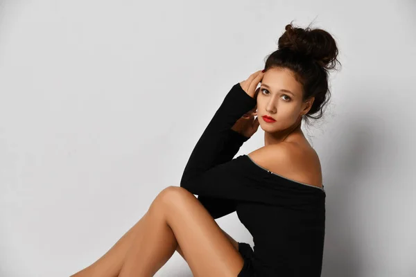 Portrait of gorgeous woman in stylish black tight dress sitting sideways to us with her legs tucked up and looking at us — 스톡 사진